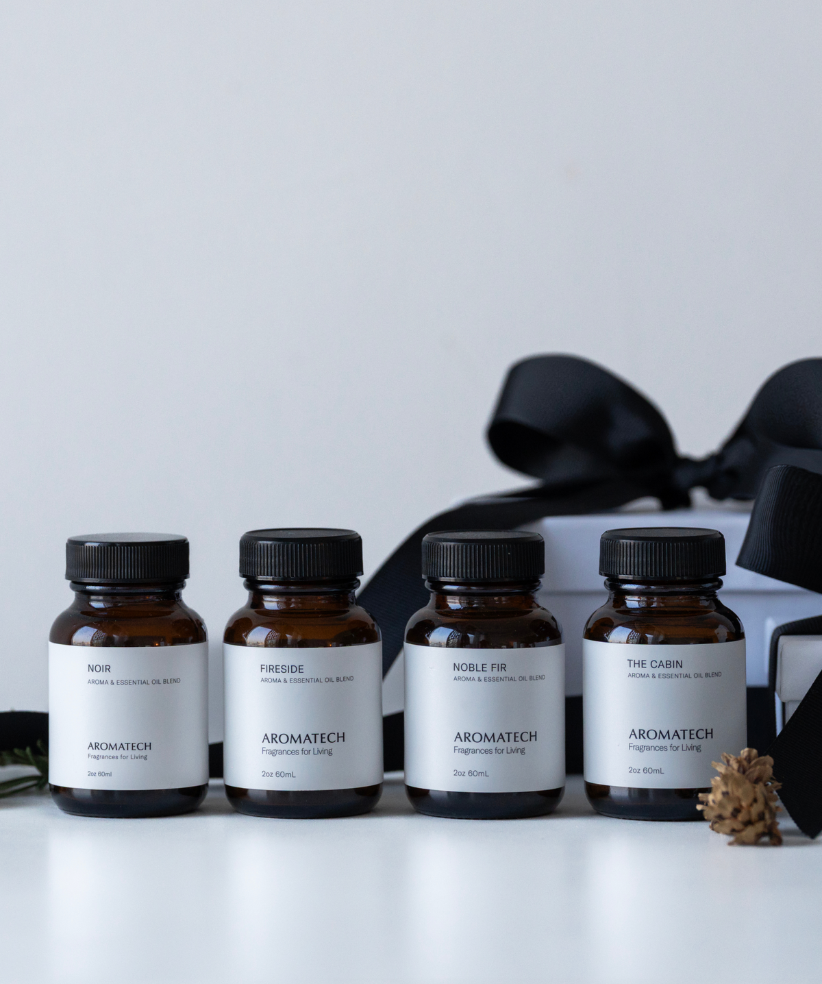 Four amber glass perfume bottles with white labels and black caps are lined up on a white surface. Behind them is a white box with a large black ribbon, and a small pinecone sits in front of the bottles.